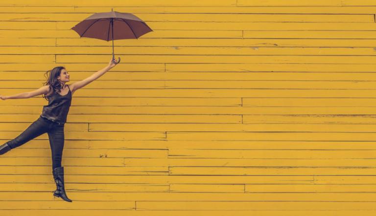 Das Foto zeigt eine in die Luft springende Frau mit einem Regeneschirm vor einer gelben Wand.
