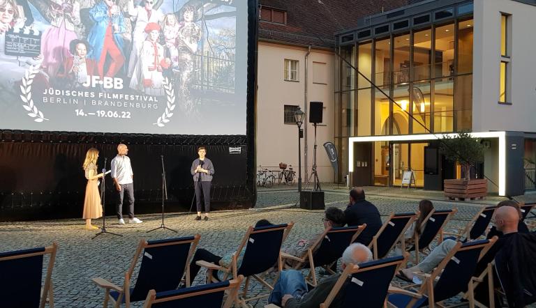 Mehrere Menschen sitzen in Liegestühlen und wohnen einem Open-Air Kino bei.