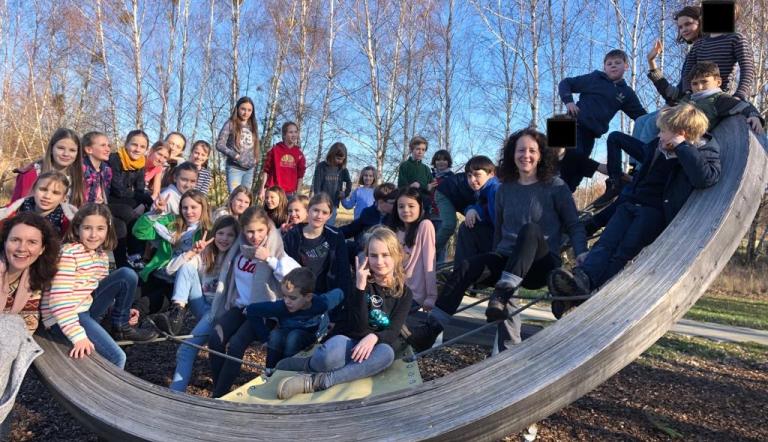 Hier ist eine sehr große Kinderschar und zwei erwachsene Frauen auf einem großen rund geschwungenem Klettergerät vor Bäumen zu sehen. Es handelt sich um das Streichorchester Die Saitentänzer der Städtischen Musikschule Potsdam "Johann Sebastian Bach"