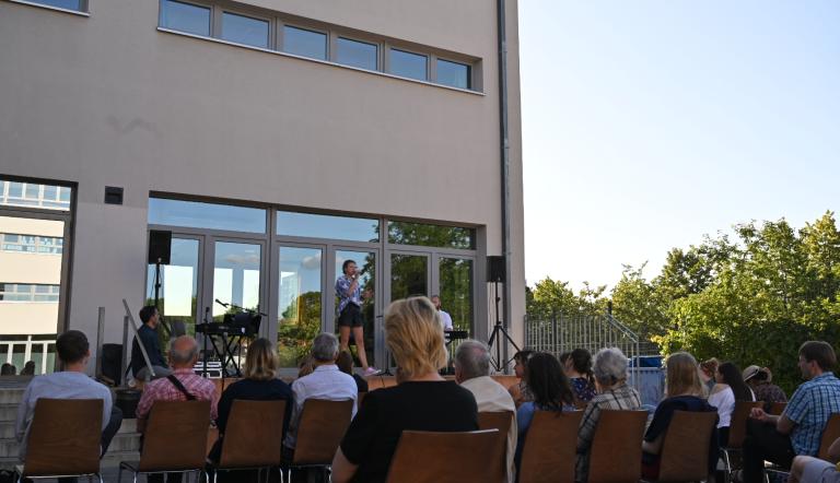 Hier erscheint ein Bild mit einer improvisierten Schulhofbühne. Eine Sängerin wird vom Keyboard begleitet und einige Angehörige bilden eine Publikum. SIe sitzen locker verteilt auf Stühlen und hören zu.