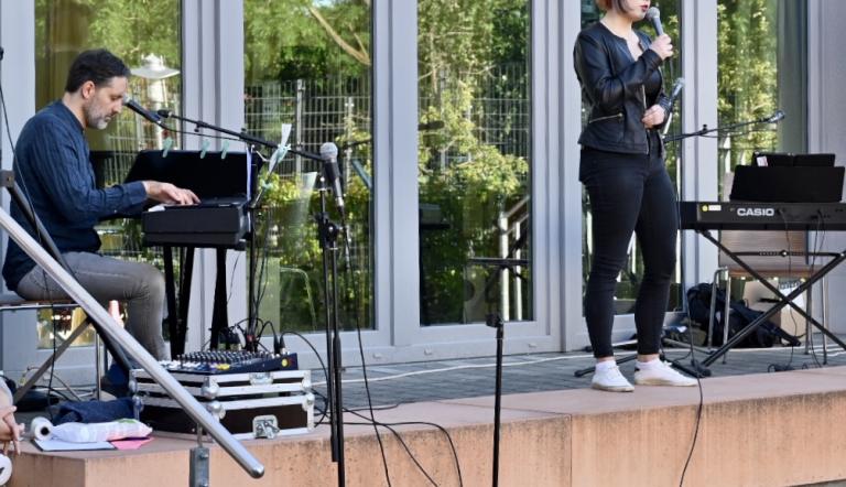Hier erscheint ein Bild mit einer improvisierten Schulhofbühne. Eine Sängerin wird von ihrem Lehrer am Keyboard begleitet.