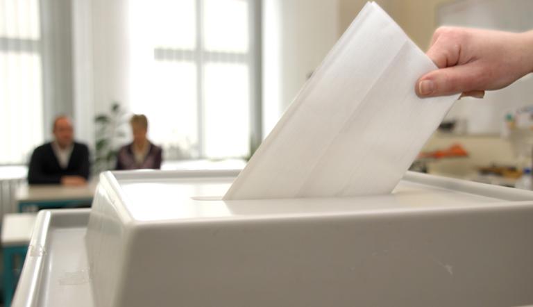 Wahlurne. Wahlurne im Vordergrund mit Hand die Wahlschein einsteckt und zwei Personen unscharf im Hinterrund