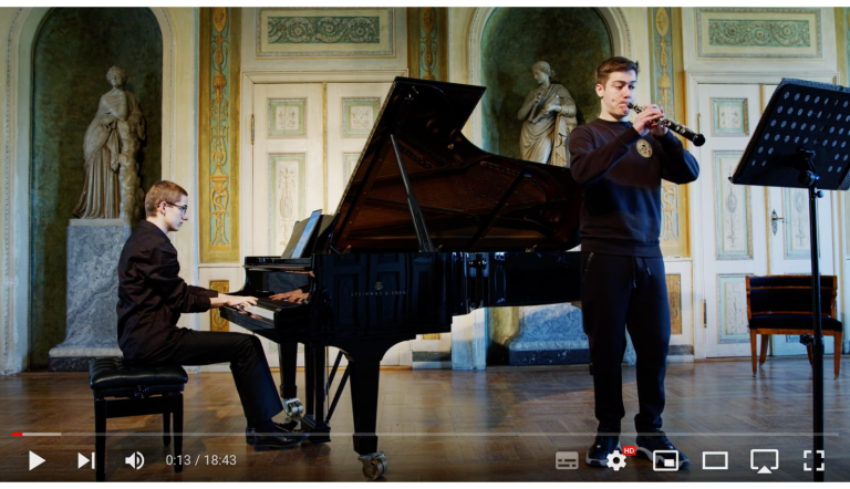 Im Saal eines Schlosses oder Palais, musizieren zwei schwarz gekleidete junge Männer an Flügel und Oboe. Die Anmutung ist sehr edel.