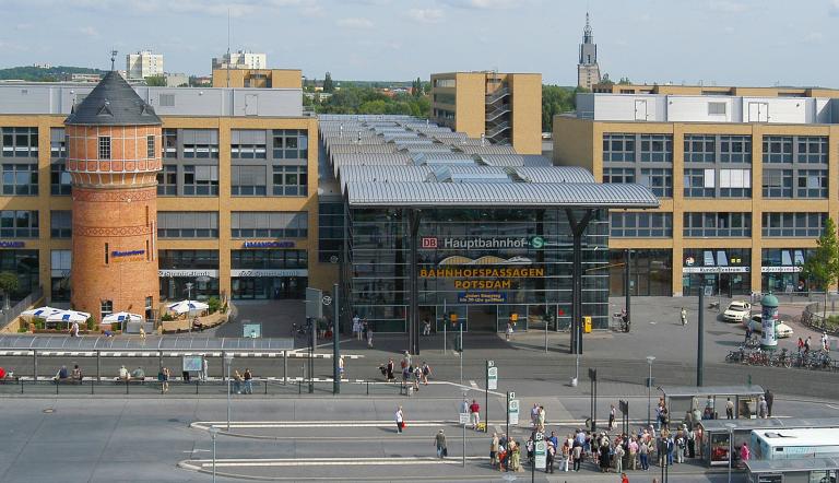 Das Bild zeigt einen Teil des Potsdamer Hauptbahnhofes am Bahnhofsvorplatz mit Bus- und Straßenbahnsteigen. Blickfang ist der große gläserne Eingang mit dem markanten wellenförmigen Dach neben dem ehemaligen Wasserturm.
