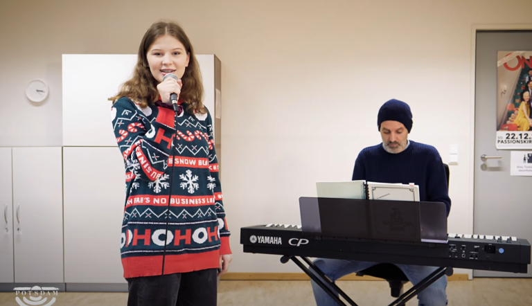 Eine jugendliche Musikschülerin singt mit Mikrophon ein Weihnachtslied - begleitet wird sie von einem Pianisten am Digitalpiano. Das ganze findet in einem hellen und modernen Unterrichtsraum der Städtischen Musikschule im Haus 2 Am Stern statt