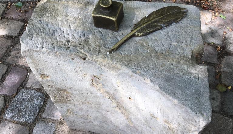 Das Foto zeigt ein bronzenes Tintenfass mit Feder  auf einem Stein-Fragmenten des ehemaligen Stadtschlosses.
