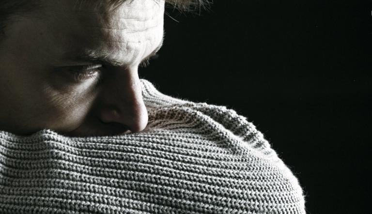 Slider: Seelische Gesundheit Das Schwarz-Weiß-Foto zeigt als Beispiel für eine traurige, depresive Stimmung einen jungen Mann, der seinen Kopf in die Armbeuge seines Pullovers drückt und selbstverloren in sich und in die Welt schaut.