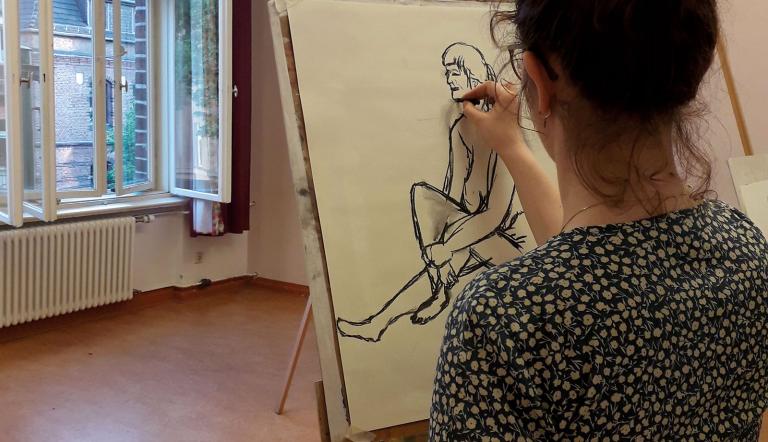 Eine Person steht mit dem Rücken zur Kamera, vor sich eine Staffelei stehend. Die Person zeichnet mit schwarzer Kohle eine unbekleidete Frau., Foto: Christoph Knäbich