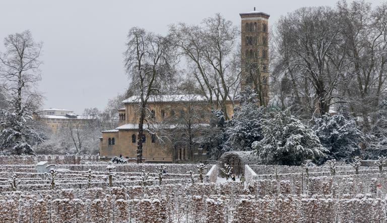 Verschneiter Park mit Hecken und kahlen Bäumen, rechts eine Kirche mit separat stehendem Kirchturm unter grauem Himmel., Foto: André Stiebitz, Lizenz: PMSG
