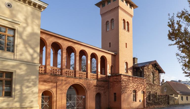 Rötlich verputzte dreibogige Toreinfahrt mit Arkadengang und Turm neben hellem Gebäude vor blauem Himmel., Foto: Sophie Soike, Lizenz: PMSG