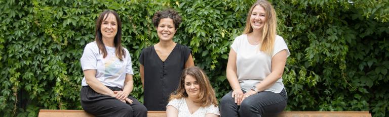 Die Beauftragten des Büros für Chacengleichheit und Vielfalt sitzen auf einer braunen Bank,  im Hintergrund ist eine grüne Hecke zu sehen (von links nach rechts: Stefanie Buhr, Dr. Tina Denninger, Claudia Sprengel, Dr. Amanda Palenberg)