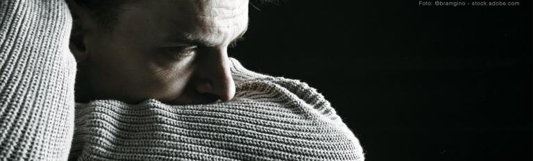 Das Schwarz-Weiß-Foto zeigt als Beispiel für eine traurige, depresive Stimmung einen jungen Mann, der seinen Kopf in die Armbeuge seines Pullovers drückt und selbstverloren in sich und in die Welt schaut.
