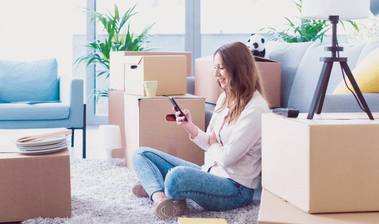 Zwischen Umzugskartons in einer ein wenig eingerichteten Wohnung sitzt eine lächelnde Frau mit ihrem Smartphone in der Hand.